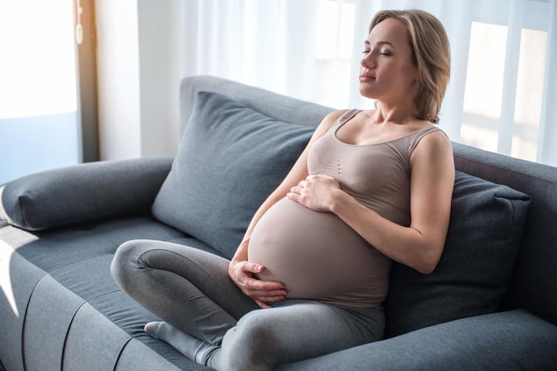 Femme enceinte détendue sur un canapé, les yeux fermés, les mains posées sur son ventre